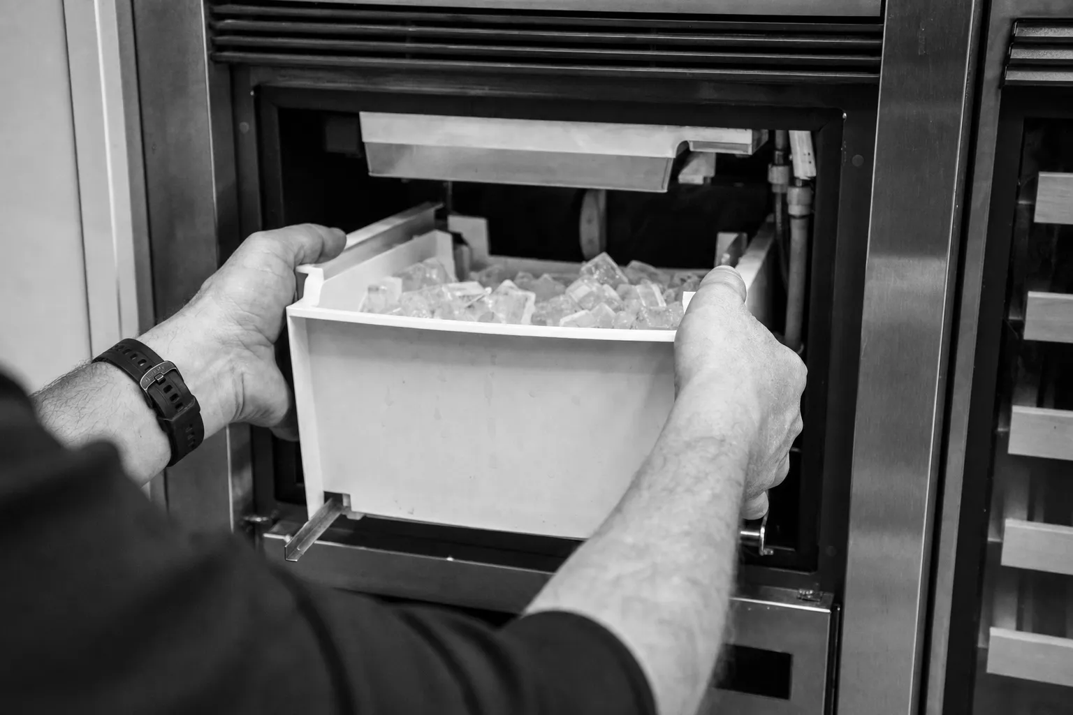 Black-and-white shot of hands lifting a full ice bin out of a built-in ice maker